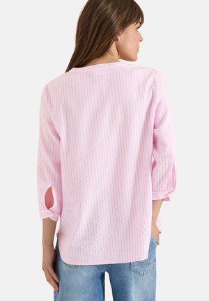 Mujer que lleva una camisa de manga larga con rayas rosa claro y las mangas remangadas, y vaqueros azules, de pie con la espalda hacia la cámara.