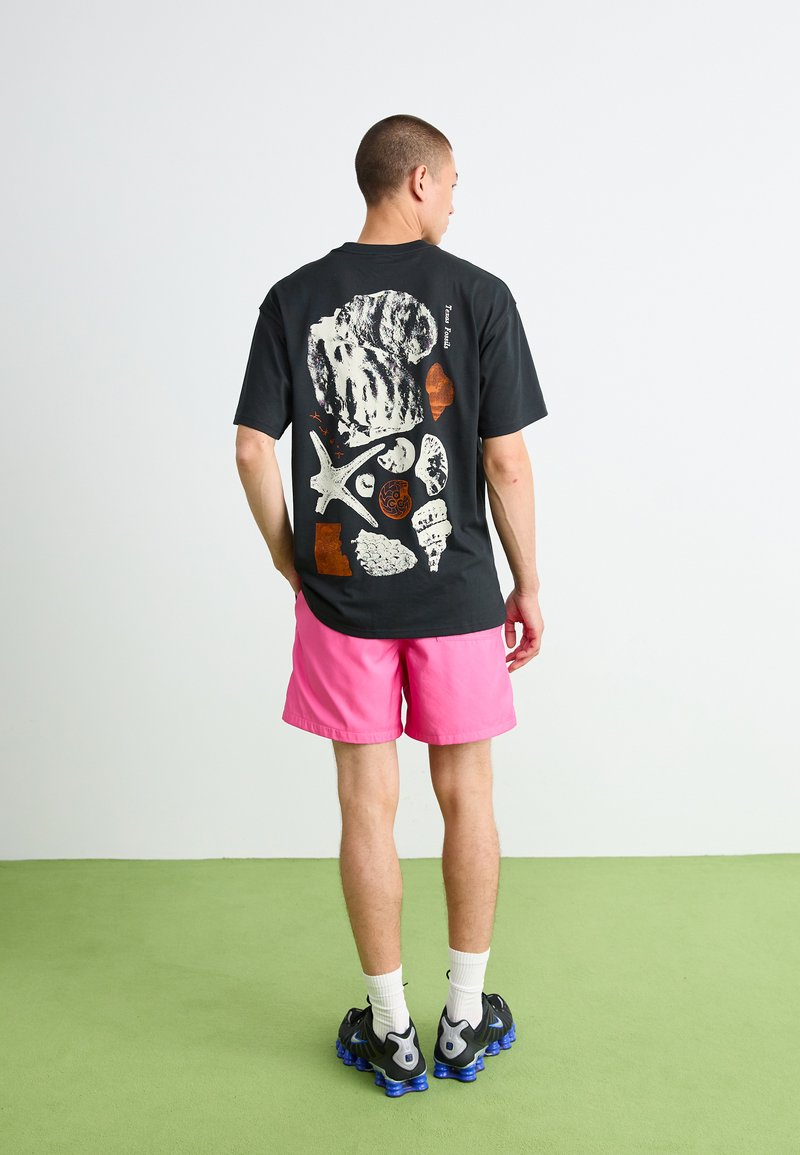T-shirt noire avec un design graphique coloré représentant des coquillages et des étoiles de mer au dos, associé à des shorts roses et des baskets noires. Sol vert.