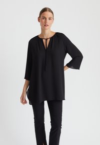 Schwarze Tunika mit Drei-Viertel-Ärmeln, Rundhalsausschnitt und Bindedetail. Glattes Material mit leicht durchscheinendem Saum und lockerer Passform.