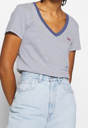 Personne portant un T-shirt à rayures bleues et blanches avec un col en V, un petit logo rouge, et un jean taille haute bleu clair.