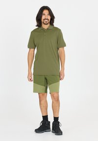 Olivgrön polo med krage, kort ärm och subtil logotyp; matchas med gröna shorts med färgspräcklig design. Svarta vandringskängor.