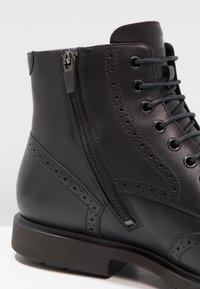 Bota de cuero negro con diseño de brogue, cordones en la parte delantera, cremallera lateral y suela plana con un ligero tacón. Textura suave con detalles de costura.