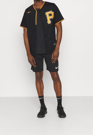 Muž ve černém baseballovém dresu Pittsburgh Pirates se žlutým lemováním, černých šortkách Nike, černých ponožkách Nike a černých teniskách stojí opřený o bílou stěnu.