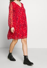 Rotes Blumenkleid mit langen transparenten Ärmeln, V-Ausschnitt und lockerer Passform. Kombiniert mit schwarzen Knöchelstiefeln mit Reißverschluss und dicken Sohlen.