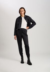 Black cropped jacket with snap buttons, worn over a white top and black jogger pants with zippered pockets, paired with black ankle boots.