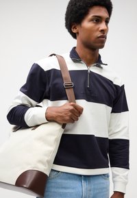 Felpa con collo zip a righe blu e bianche, con polsini a coste, abbinata a una borsa tote beige con accenti in pelle marrone.