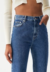 High-waisted blue denim jeans with a faded finish, five pockets, and gold hardware. The fabric texture is smooth with a slight sheen.