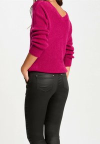 Frau in einem leuchtend pinken, langärmeligen Strickpullover und engen schwarzen Hosen, mit den Händen in den vorderen Taschen, von hinten gezeigt.