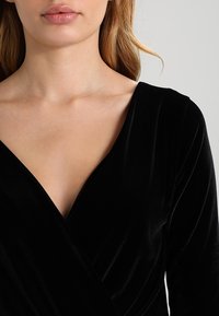 Top de terciopelo negro con un diseño de escote en V profundo, que presenta mangas largas y una textura suave y lisa. Sin patrones visibles ni detalles de herrajes.