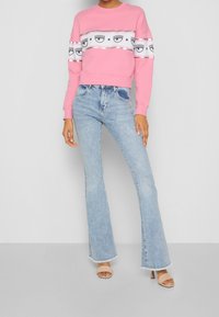 Sudadera rosa con patrón gráfico y acentos plateados, combinada con vaqueros acampanados azul claro y tacones abiertos en tono nude.