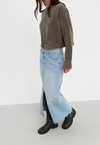 Pull marron court avec ourlet côtelé, jupe en jean bleu clair avec fente frontale, et bottines noires ornées de clous décoratifs.