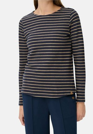 Femme portant un chemisier bleu marine à manches longues avec de fines rayures horizontales beige et un pantalon bleu foncé, debout devant un fond uni.