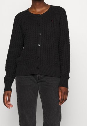 Personne portant un cardigan noir boutonné en maille torsadée à manches longues et un jean noir, debout devant un fond clair uni.
