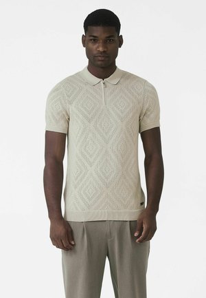 Beežikiudu lühikeste varrukatega polo kampsun teemantmustriga, krae ja tõmblukuga. Sobitatud helehallide pükstega, mis on lõdva lõikega.