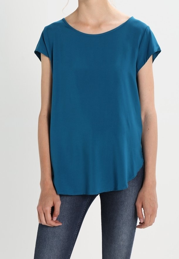 Femme portant un chemisier turquoise à manches courtes et un jean bleu foncé, debout devant un fond clair uni.