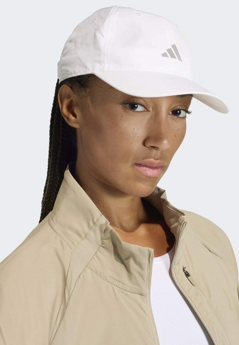 Gorra de béisbol blanca con visera curvada y logotipo gris, combinada con una chaqueta gris claro con cremallera que presenta un cuello texturizado y un bolsillo lateral.