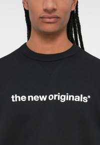 Sweatshirt preto com gola canelada, apresentando o texto branco &quot;the new originals®&quot; atravessando o peito. Textura suave.