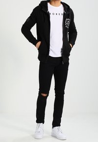 Svart zip-up hoodie med fickor, vit grafisk t-shirt, svarta ripped skinny jeans och vita sneakers. Avslappnad urban outfit.