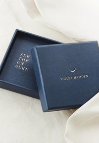 Dunkelblaues quadratisches Kästchen mit einer strukturierten Oberfläche. Innen steht in goldenen Buchstaben "SEE THE UN SEEN" und "VIOLET HAMDEN" mit einem Detail eines Halbmonds.