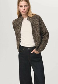 Chaqueta bomber marrón de punto con cuello alto, cierre de cremallera y puños acanalados, usada sobre una camisa clara y vaqueros oscuros.