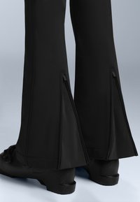 Czarne, dopasowane spodnie z ukośnymi zamkami na końcach nogawek, o gładkiej fakturze i lekko rozszerzonym kroju, zestawione z czarnym obuwiem.