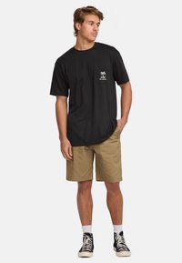 Camiseta de algodón negra con bolsillo en el pecho que presenta un gráfico de palmera, combinada con pantalones cortos caquis, calcetines blancos y zapatillas altas negras.