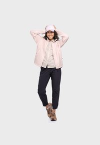 Hellrosa Fleecejacke, beiges Hemd mit Knöpfen, marineblaue Hose und hellbraune Sneaker. Model trägt einen hellrosa Hut, die Arme erhoben, und lächelt.