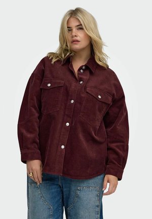 Frau mit blondem Haar, die ein weinrotes Cordhemd mit Knöpfen und blaue Jeans aus Denim mit großen Vorderaschen trägt.