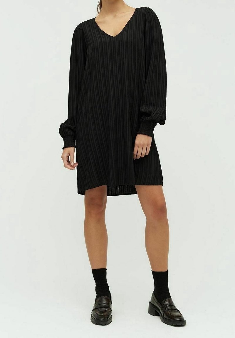 Femme portant une robe noire plissée à manches longues et longueur genou, avec des chaussettes noires et des mocassins noirs à talons, debout sur un fond blanc.