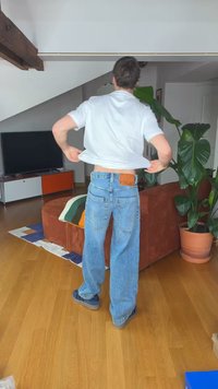 Άνδρας με μπλε τζιν και λευκό πουκάμισο σηκώνει το πουκάμισο, στέκεται σε σαλόνι με καφέ καναπέ, μεγάλο πράσινο φυτό και τηλεόραση στο φόντο.