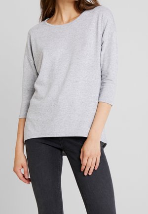 Femme portant un haut gris clair à manches longues avec un col rond et un jean noir ajusté, sur un fond neutre.