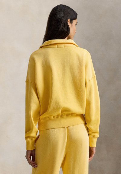 Κίτρινο πουλόβερ με άνετη εφαρμογή, ραβδωτή υφή και ψηλό γιακά. Διαθέτει ελαστική ζώνη στο κάτω μέρος και φαρδιά μανίκια, συνδυασμένο με παντελόνι στο ίδιο χρώμα.
