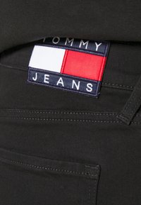 Černé džíny s texturovaným povrchem, s obdélníkovou logem nášivkou v červené, bílé a modré barvě se štítkem "TOMMY JEANS" na pásku.