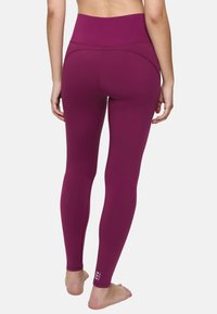 Femme debout pieds nus portant un legging violet taille haute, vue de dos sur un fond blanc uni.