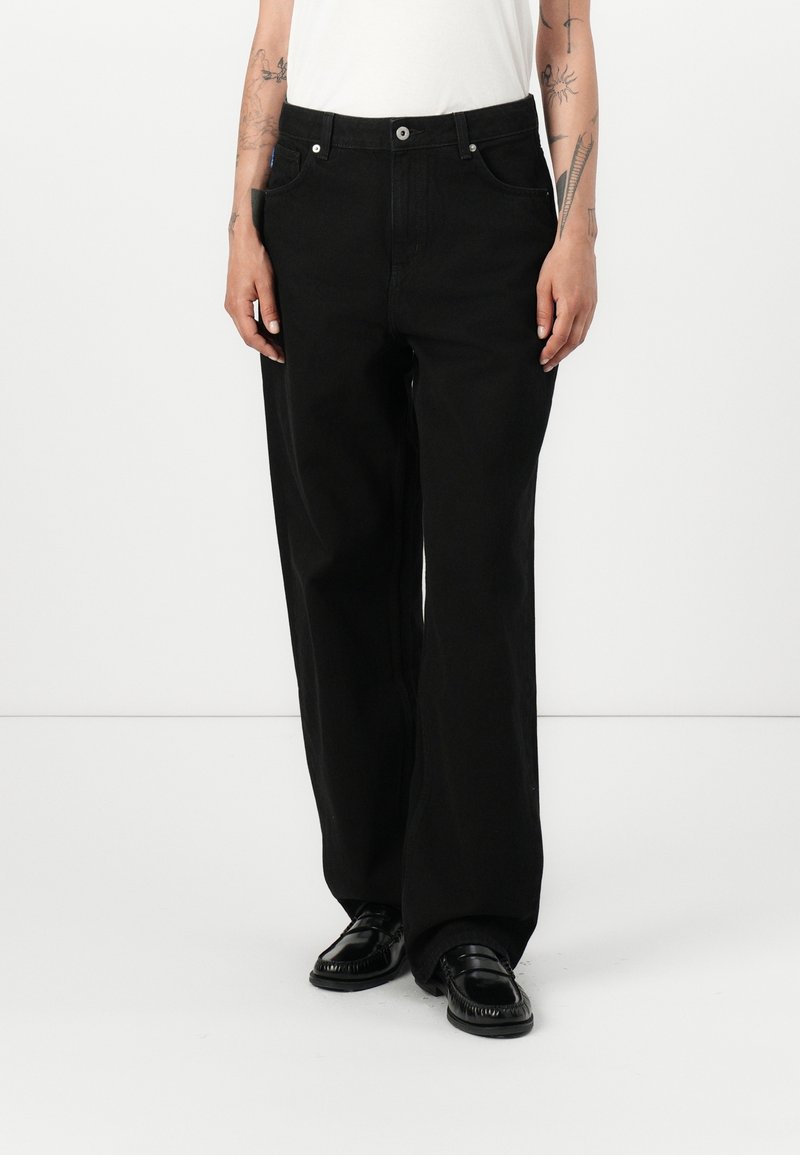 Personne portant un jean noir à jambes larges, des mocassins noirs et un haut blanc sans manches, debout avec les mains détendues le long du corps.