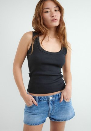 Mladá žena s dlhými vlasmi, oblečená v čierny top bez rukávov a modré rifľové šortky, stojí s rukami v vreckách.