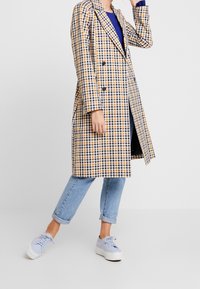 Doppelreihiger Trenchcoat in Gelb, Marineblau und Weiß mit Karomuster, mit geknöpftem Kragen, langen Ärmeln und lockerer Passform. Getragen mit hellblauen Jeans und Turnschuhen.