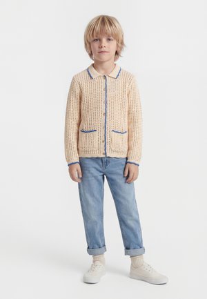 Ξανθό παιδί που στέκεται φορώντας ένα εκρού πλεκτό ζακέτα με μπλε περιθώριο, ανοιχτό μπλε τζιν παντελόνι γυρισμένο, μπεζ κάλτσες και λευκά αθλητικά παπούτσια μπροστά σε λευκό φόντο.