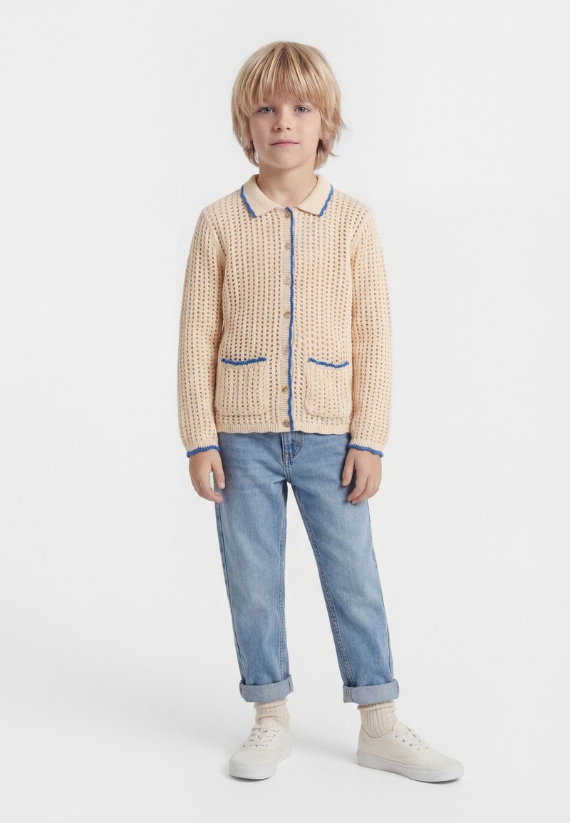 Blond dieťa stojace a oblečené v krémovom pletenom kardigáne s modrým lemom, svetlomodrých vyhrnutých džínsach, béžových ponožkách a bielych teniskách na bielom pozadí.