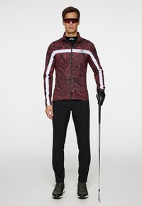 Röd mönstrad atletisk jacka med vita detaljer, svarta byxor och svarta handskar. Modell som håller en golfklubba och bär solglasögon.