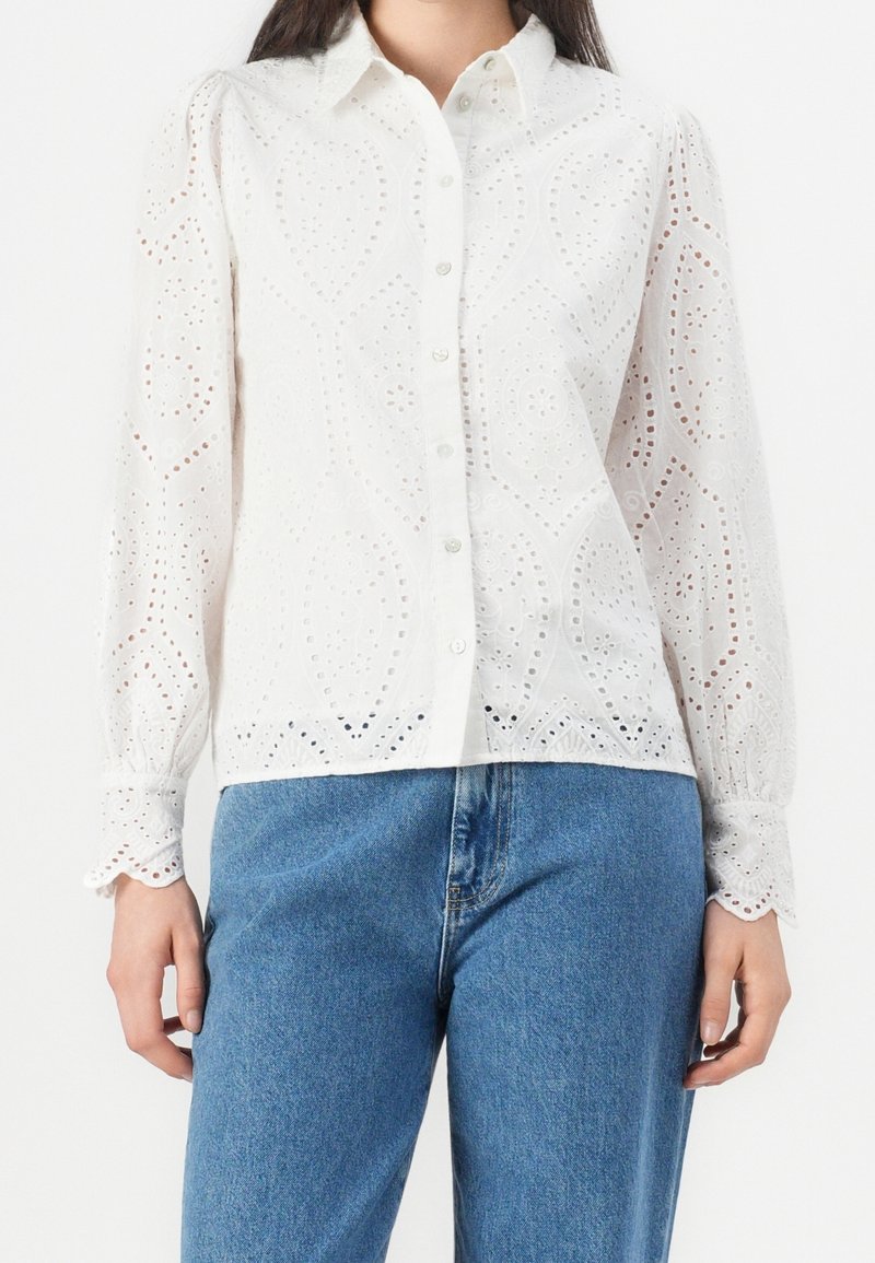 Vit långärmad blus i ögla med knappar framtill och vågformade ärmslut, som bärs tillsammans med blå denimjeans.