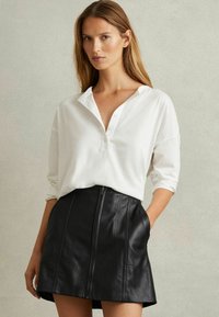 Camiseta henley de manga larga blanca con tapeta y falda corta de cuero negra con cremallera frontal y bolsillos laterales, con una textura suave.