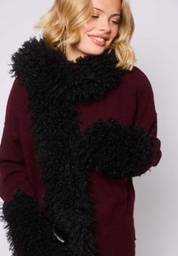 Donna bionda con un maglione bordeaux, avvolta in una lunga sciarpa nera riccia e texturizzata, che sorride e guarda verso il basso.