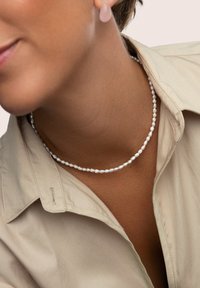 Die Perlenkette besteht aus kleinen, unregelmäßig geformten weißen Perlen, die aneinandergereiht sind und zu einem beigen Kragenhemd mit glatter Textur passen.