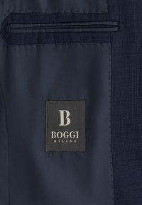 Interno di una giacca da abito blu scuro che mostra la tasca interna e un'etichetta in tessuto nero con la scritta "B BOGGI MILANO" in bianco.
