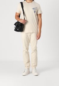 Béžové bavlněné tričko s logem, krémové kalhoty a bílé tenisky. Černá crossbody taška připevněná na popruhu, se zipem a přezkou.