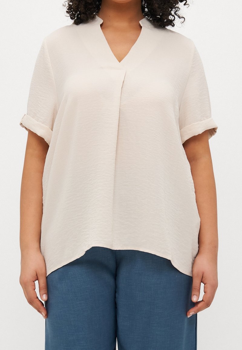 Blouse beige à manches courtes avec un col en V et un pli devant. Tissu doux et texturé ; ourlet légèrement arrondi. Associée à un pantalon bleu.