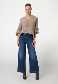 Beige kabelgebreide trui met een V-hals, gecombineerd met hoogwagende, brede blauwe spijkerbroeken. Model draagt donkere enkellaarzen.