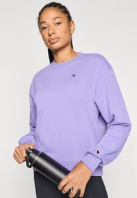 Μωβ φούτερ με μακριά μανίκια, στρογγυλή λαιμόκοψη και λογότυπο στο στήθος. Διαθέτει ριμπ μανσέτα και τελείωμα. Κρατά μαύρο παγούρι νερού.