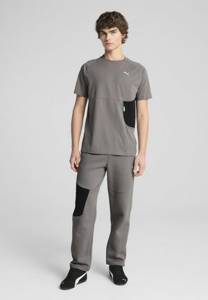 Conjunto deportivo gris que presenta una camiseta de cuello redondo con un bolsillo negro y cremallera, combinada con pantalones a juego que tienen acentos y bolsillos negros.